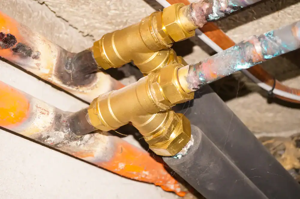 Close-up of copper pipes joined by brass fittings under a ceiling, with some pipes showing corrosion, rust, and mineral buildup. The pipes are aligned horizontally and run parallel to each other.