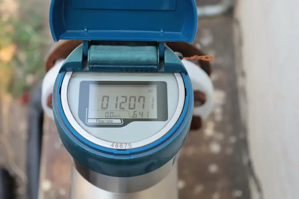A close-up of a digital water meter displaying a reading of 012071 cubic meters. The device has a blue cover, and the meter is installed outdoors on a water pipe.