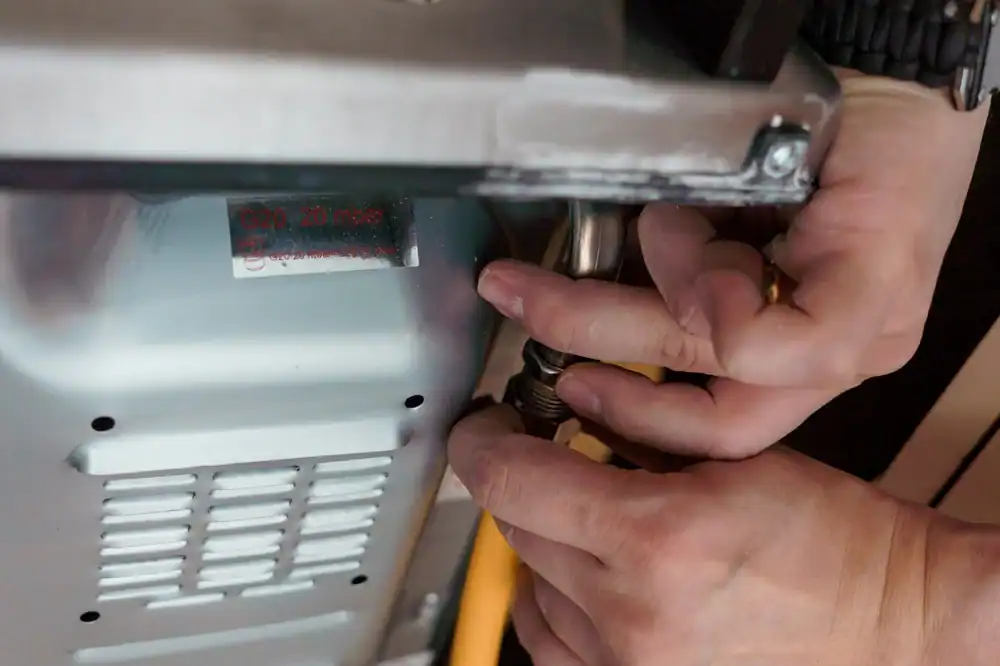 A close-up of hands tightening a gas hose connection on the back of a metal appliance, possibly a stove or oven, with a yellow hose visible.