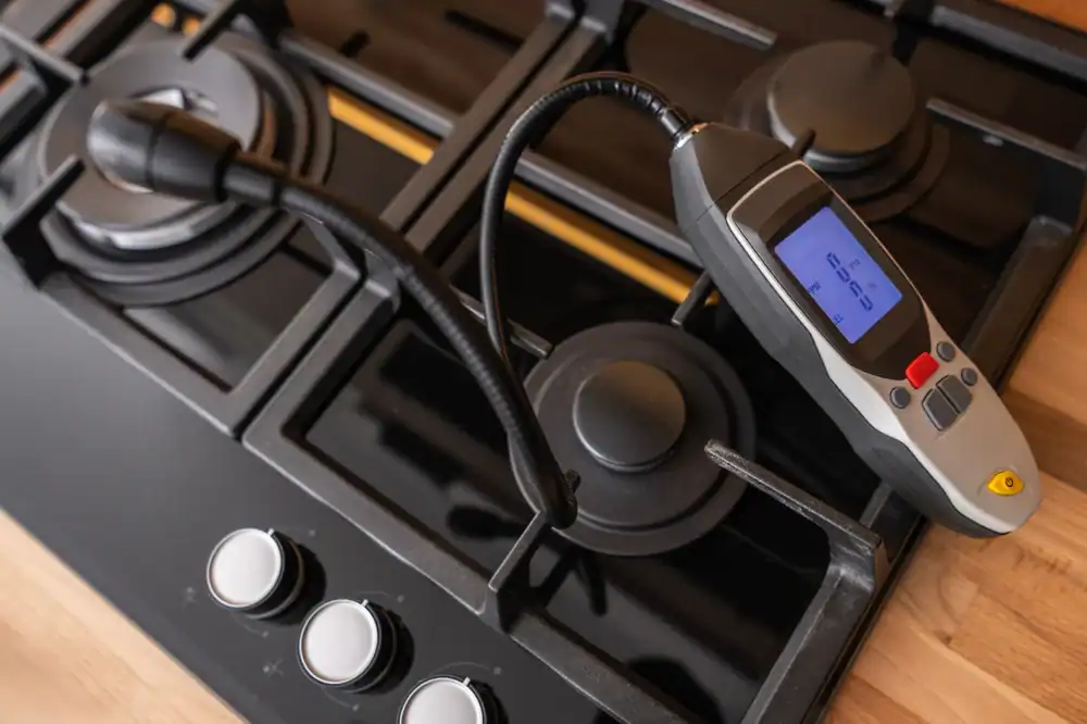 A gas leak detector with a digital display is placed on the burners of a black gas stove, with control knobs visible in the foreground.