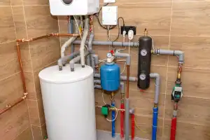 Home boiler room with a white water heater, blue expansion tank, various pipes, valves, pressure gauges, and control devices mounted on wood-patterned tiled walls.