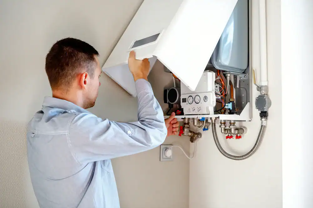 A man in a light blue shirt inspects and repairs a wall-mounted gas boiler, with its front panel open, exposing various pipes, wires, and components inside.