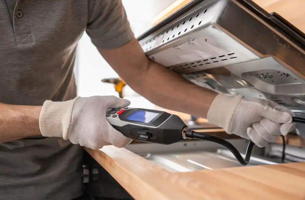 A person wearing gloves inspects the underside of a dishwasher or oven using a handheld inspection camera with a flexible probe and a screen.