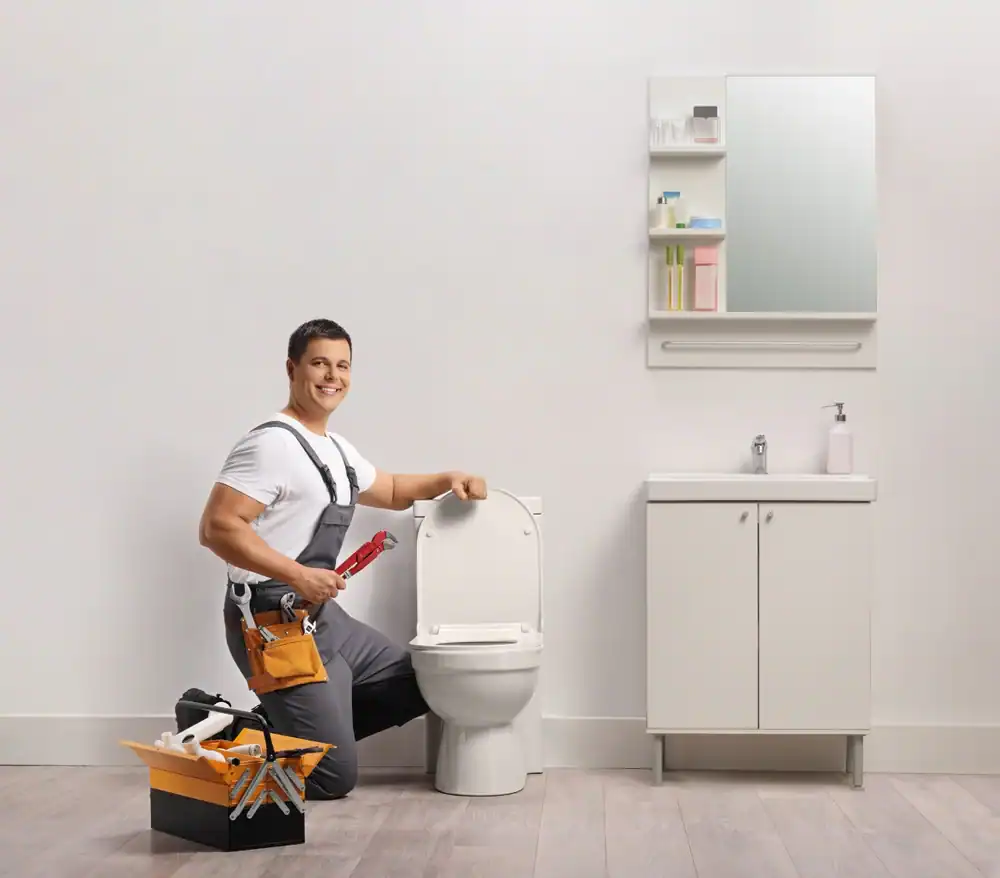 A smiling plumber in overalls kneels by a toilet holding a red wrench in a modern bathroom. A toolbox with tools sits nearby, and there is a white sink cabinet and mirror on the wall.