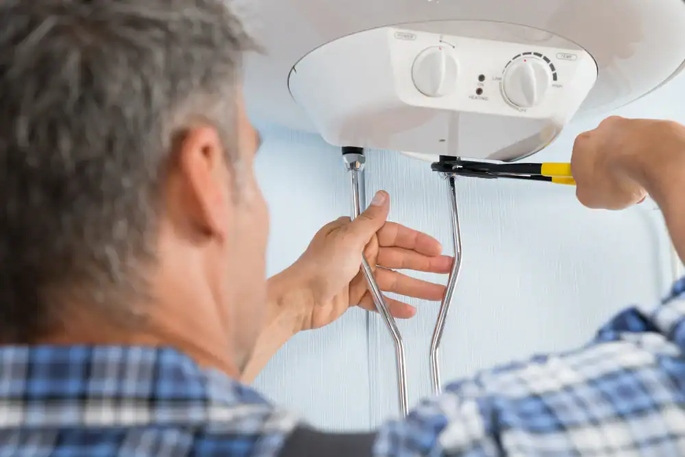A person in a plaid shirt uses a wrench to tighten pipes connected to a white water heater mounted on a wall. The focus is on the hands and tools, with the person's face out of view.