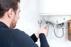 A person wearing a dark sweater uses a wrench to adjust or repair pipes connected to a white water heater on a tiled wall.