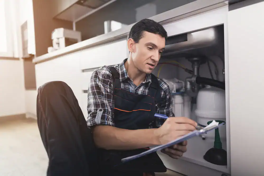 A plumber wearing overalls sits on the floor next to an open kitchen cabinet, examining pipes and writing on a clipboard. Tools and plumbing equipment are visible in the cabinet.