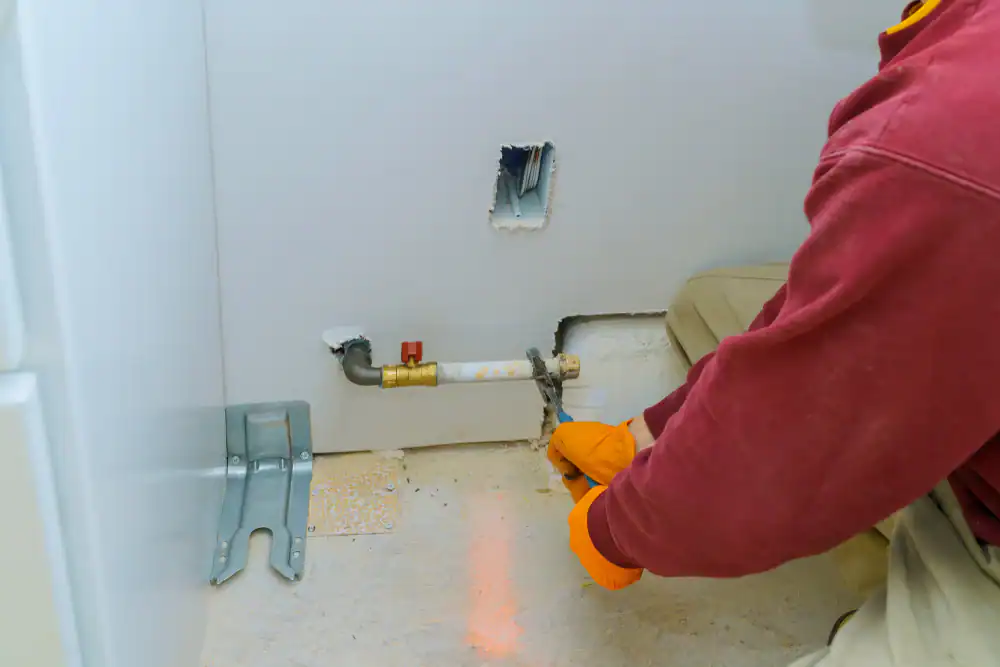 A person wearing orange gloves and a red shirt works on a gas pipe connection in a partially finished room, using a wrench near a wall with an exposed pipe and rough cutout.