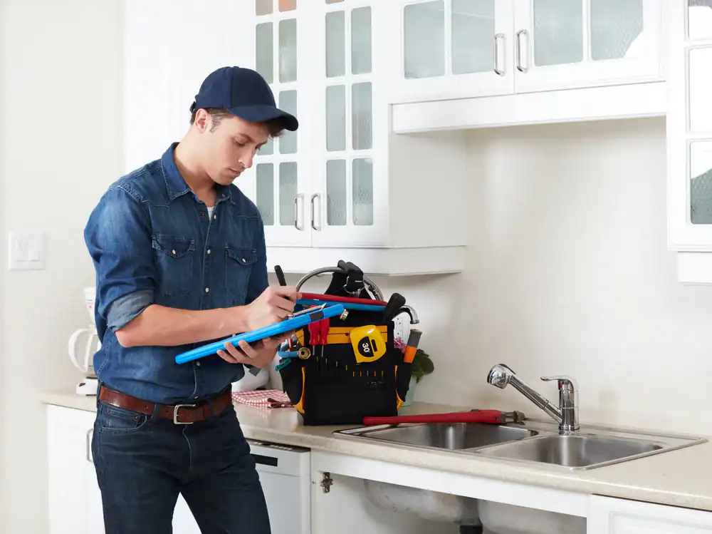 A person in a denim shirt and cap writes on a clipboard while standing in a kitchen near a sink. A tool bag with various tools sits on the counter next to them.
