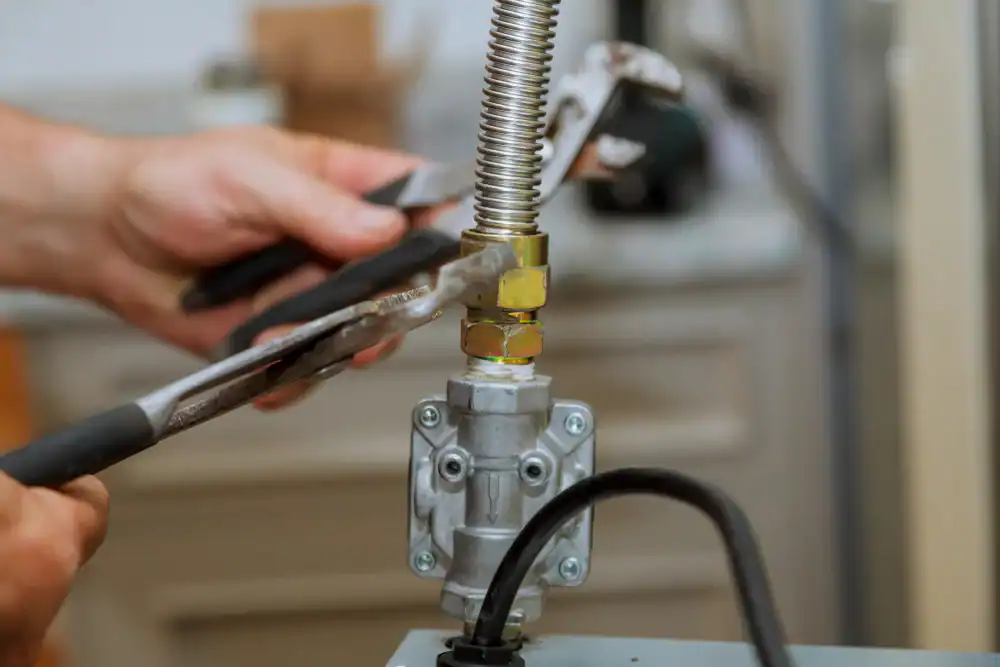 Close-up of a person using two wrenches to tighten a metal gas pipe connection, focusing on the hands and tools, with a blurred background of a kitchen or workshop.