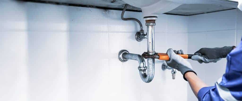 A person wearing gloves uses a wrench to repair or tighten a pipe under a sink against a white tiled wall, demonstrating the skilled service typical of a Plumbing Company New York County & Staten Island.