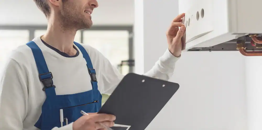 A person in blue overalls and a white shirt checks a wall-mounted control panel, holding a clipboard and pen, likely performing maintenance or inspection in a bright indoor setting.