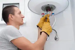 A man wearing yellow work gloves uses pliers to repair or install an electric water heater mounted on a white tiled wall.