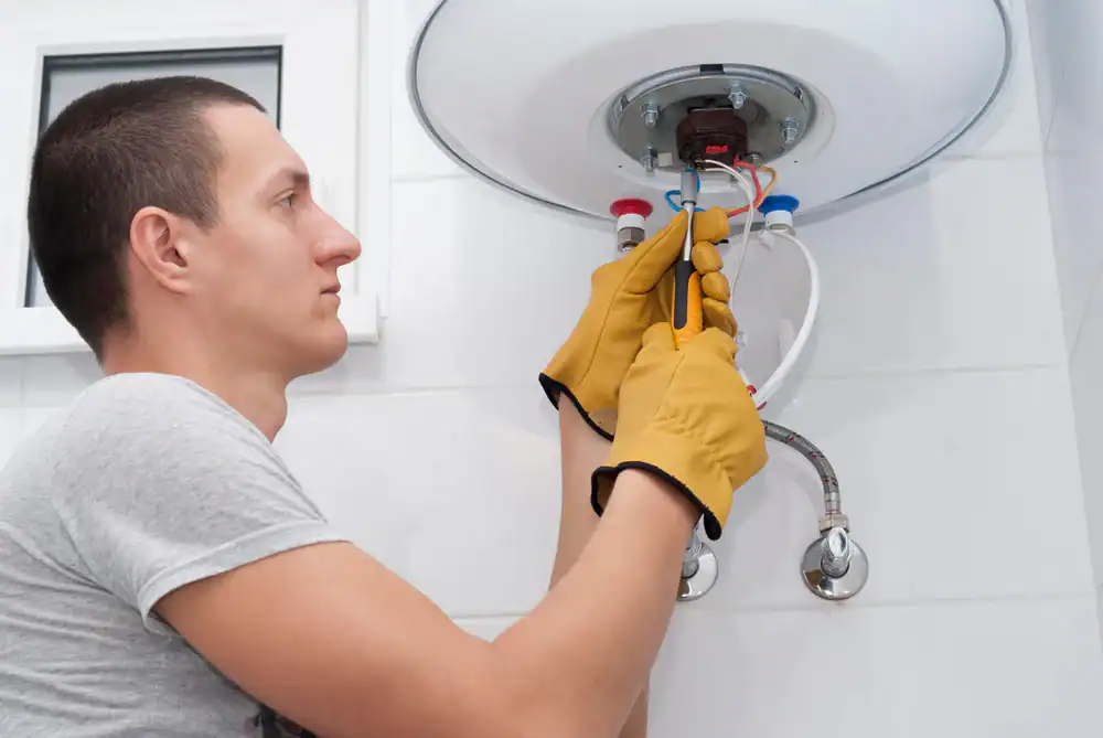 A man wearing yellow work gloves uses pliers to repair or install an electric water heater mounted on a white tiled wall.