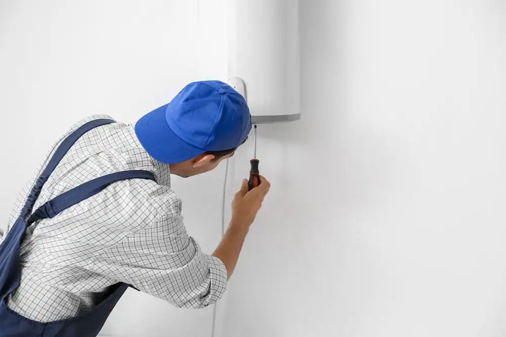 A man wearing a blue cap, checkered shirt, and overalls uses a screwdriver to repair or install a white water heater mounted on a white wall.