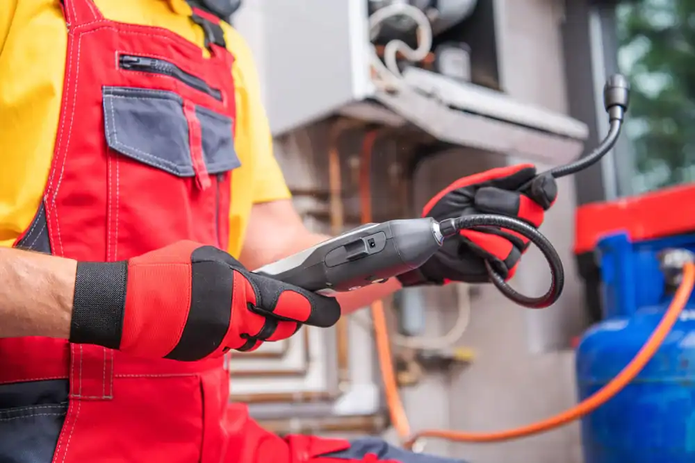 Person wearing red overalls and gloves holding a gas leak detector in front of a gas boiler, with tools and equipment visible in the background.