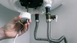 A person’s hand is installing or repairing wiring on the underside of a water heater, with metal flexible hoses and colored connectors visible.