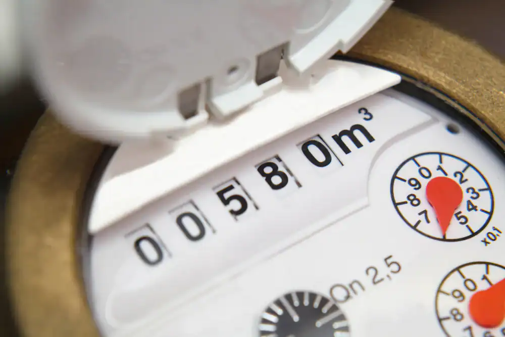 A close-up of a water meter showing the reading "00580" cubic meters, with dials and a partially open white cover visible at the top of the device.