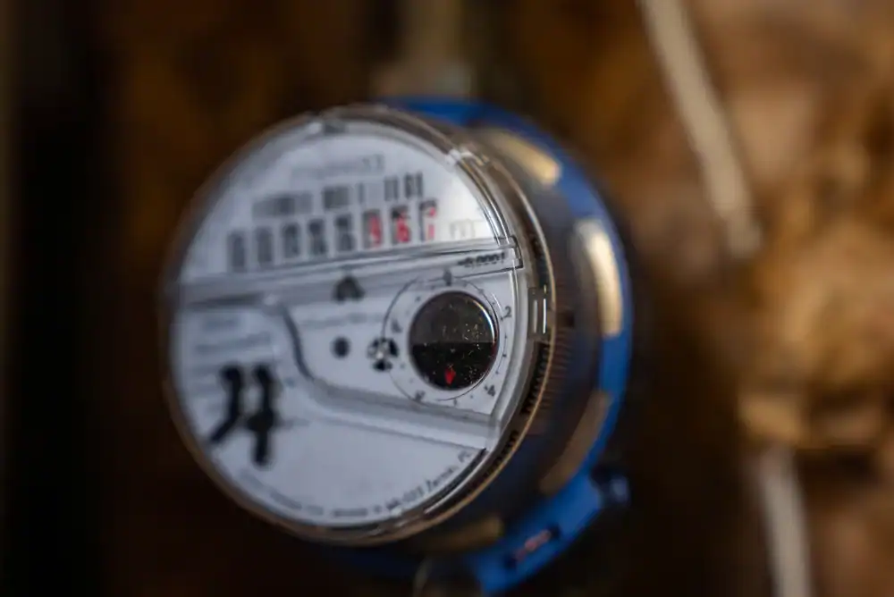 A close-up of a water meter with numbers and dials visible, mounted on a wall with pipes in the background. The focus is on the meter&rsquo;s face, showing measurement indicators.