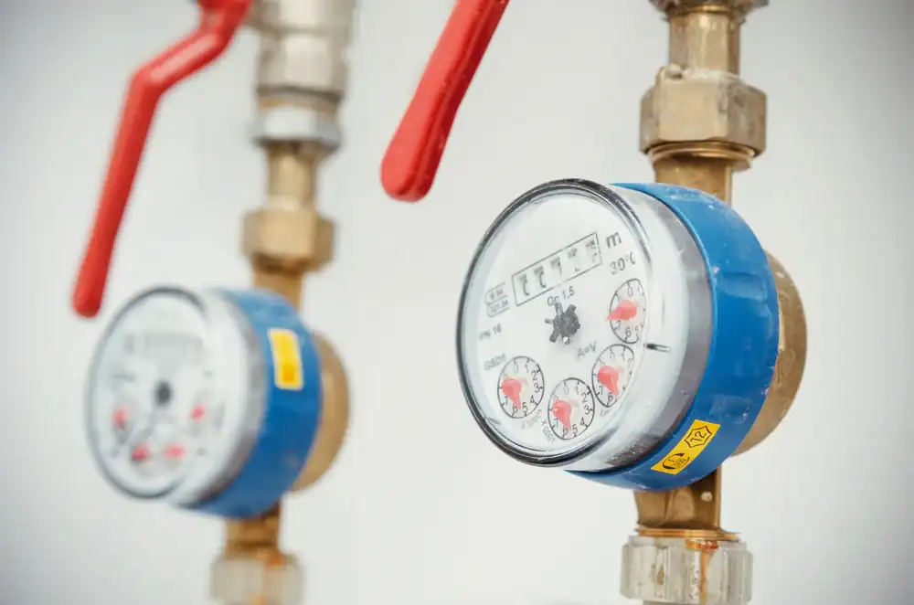 Close-up of two water meters with blue casings and mechanical dials attached to brass pipes; each has a red shutoff lever handle and yellow sticker, mounted against a white wall.