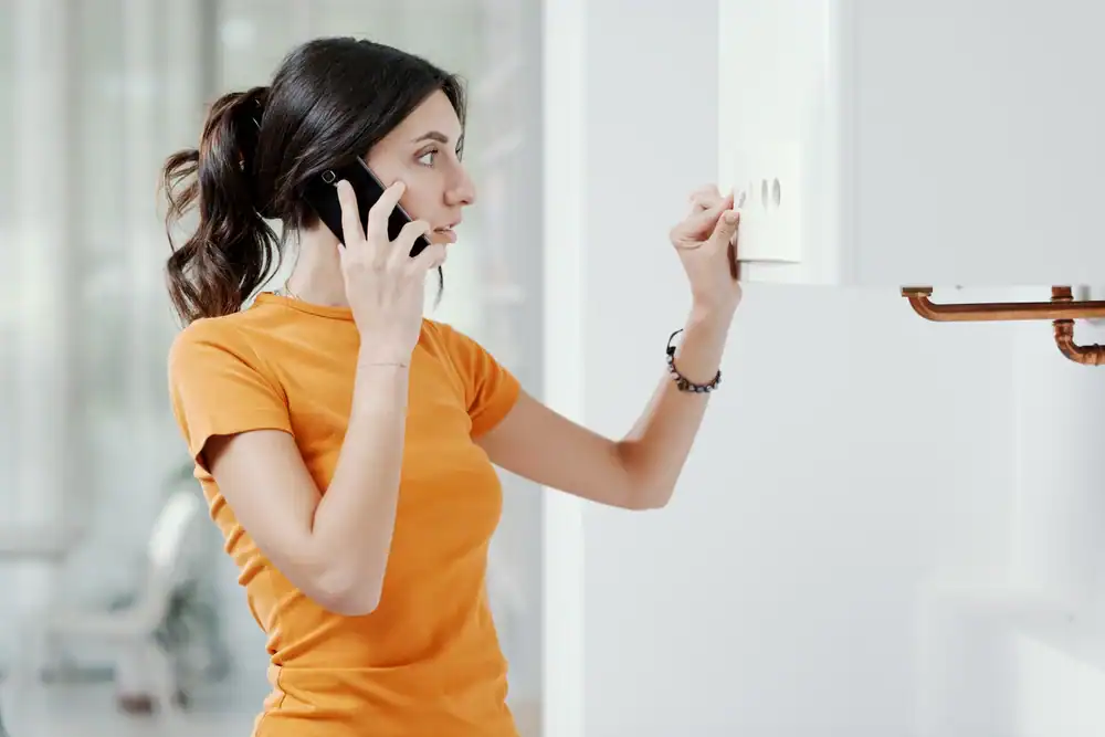 A woman in an orange shirt talks on the phone while adjusting the controls on a white wall-mounted appliance, possibly a boiler or heater, in a bright indoor setting.