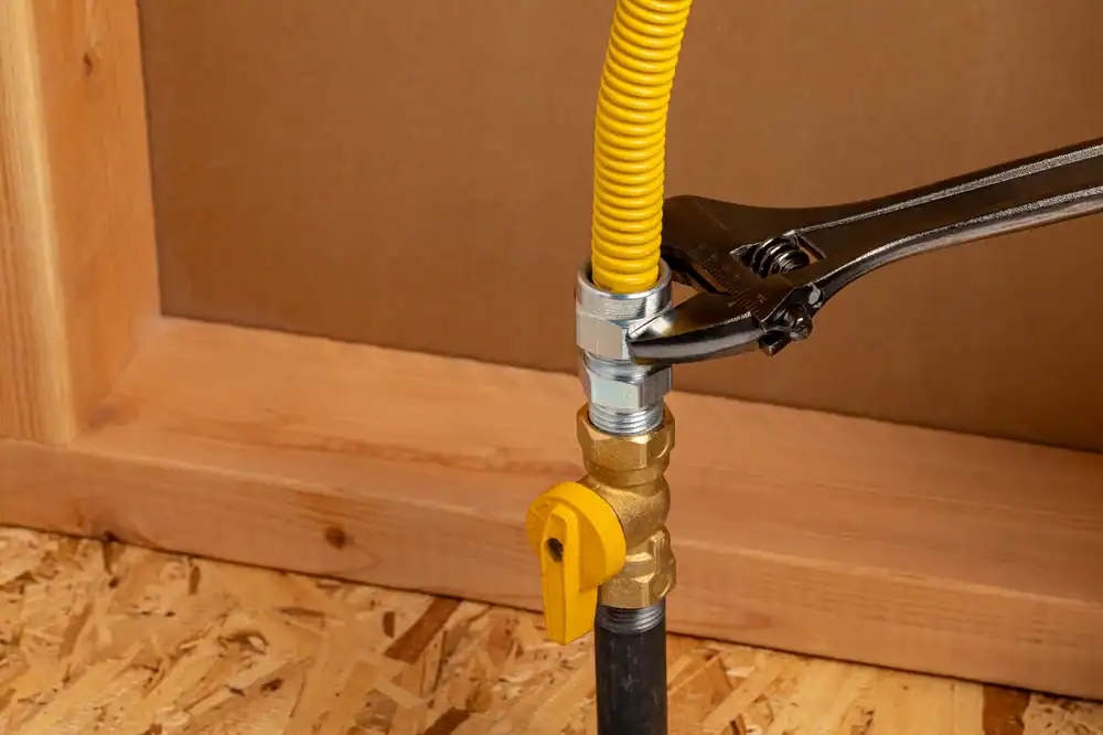 A wrench tightens a metal fitting connecting a yellow flexible gas line to a black pipe, with a brass shutoff valve attached, on a plywood surface near a wooden frame.