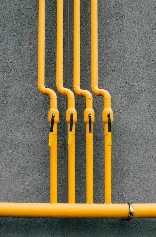 Four yellow gas pipes with shut-off valves, possibly supplying a water heater, are mounted vertically on a gray textured wall, with a horizontal pipe connecting at the bottom.