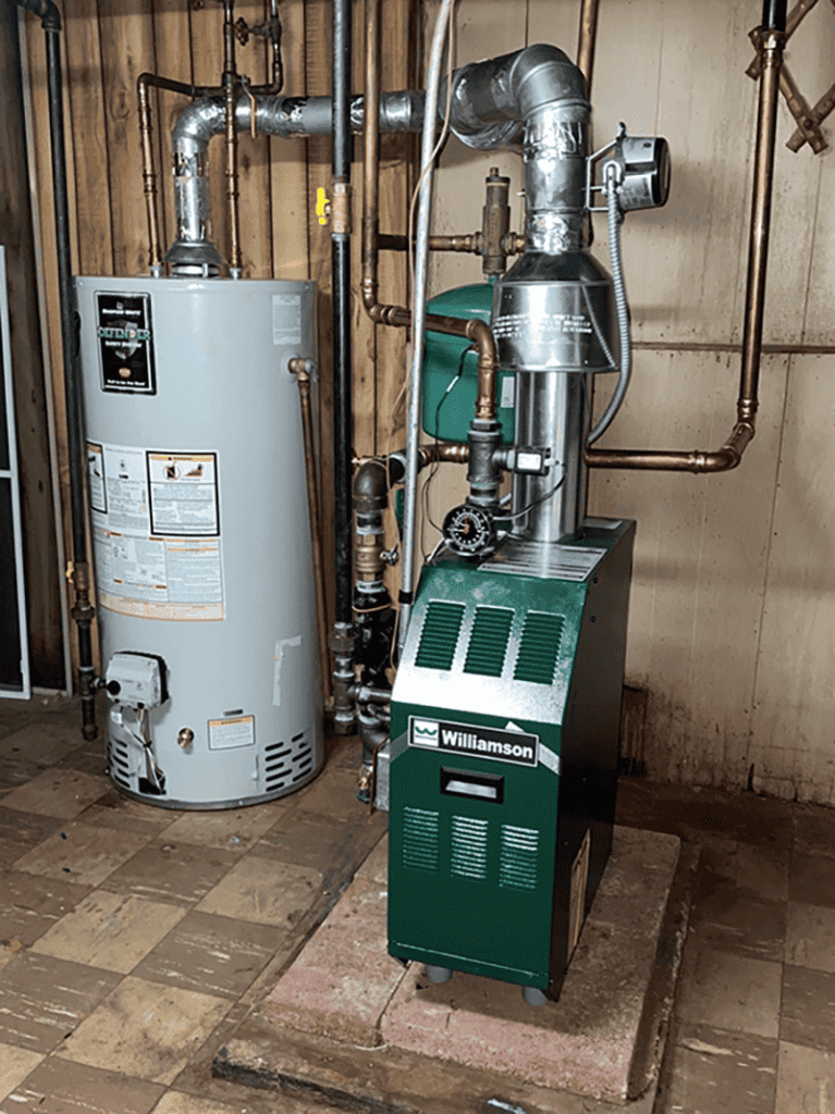A green Williamson furnace and a gray water heater are installed in a basement with exposed pipes, unfinished wooden walls, and tiled floor—expertly set up by a leading Plumbing Company in New York County & Staten Island, NY.