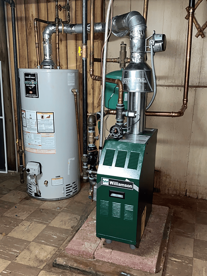 A green Williamson furnace and a gray water heater are installed in a basement with exposed pipes, unfinished wooden walls, and tiled floor&mdash;expertly set up by a leading Plumbing Company in New York County & Staten Island, NY.