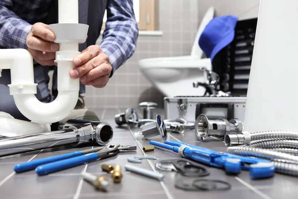 A plumber from a leading Plumbing Company New York County & Staten Island, NY repairs a white PVC pipe under a sink, surrounded by tools and plumbing parts on a tiled bathroom floor. A toilet, toolbox, and blue cap are visible in the background.