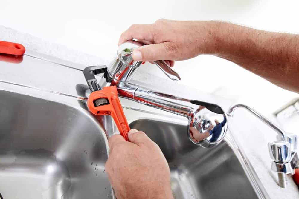 A person from a Plumbing Company New York County & Staten Island uses a red pipe wrench to tighten or fix a chrome kitchen faucet above a stainless steel sink in NY.