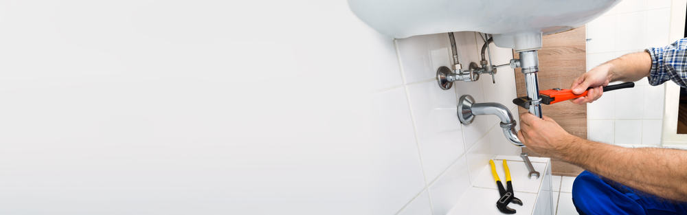 A person from a Plumbing Company in New York County & Staten Island, NY uses a wrench to fix pipes under a white bathroom sink; other tools, including pliers and a wrench, are placed on the tiled floor nearby.