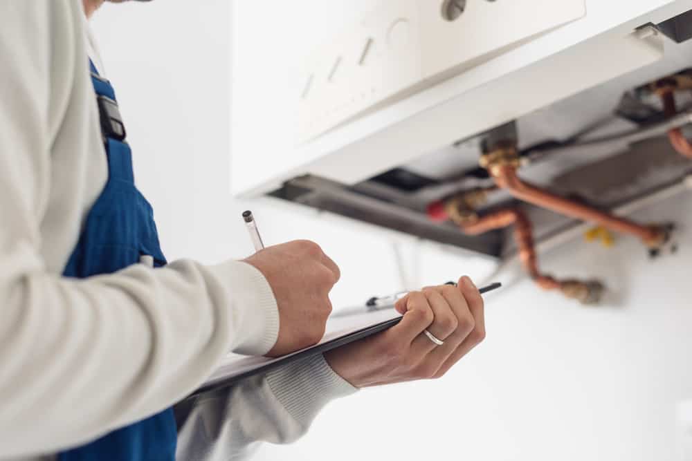 A technician in blue overalls from a leading Plumbing Company New York County & Staten Island writes on a clipboard while inspecting the pipes and components of a wall-mounted boiler or heating system in NY.