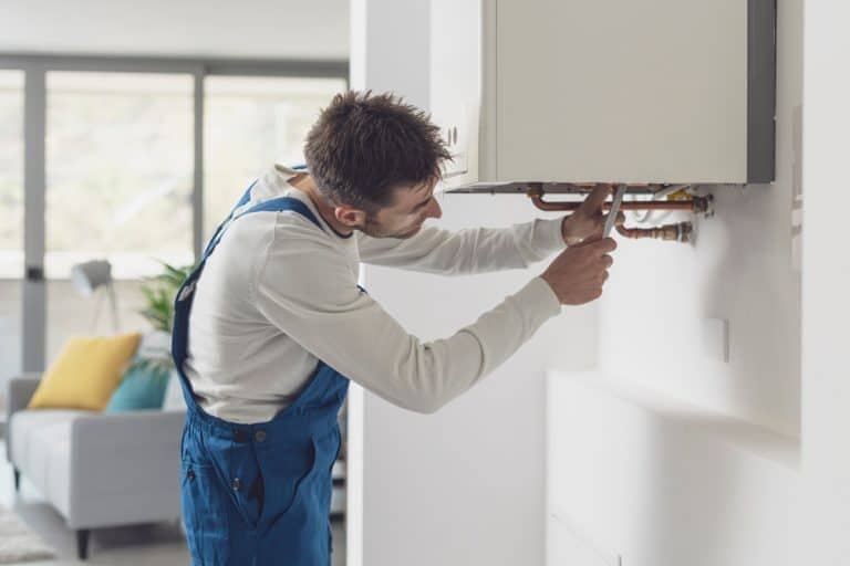 A maintenance worker in blue overalls uses a wrench to fix or install pipes under a wall-mounted boiler in a modern, bright living room&mdash;typical service from a top Plumbing Company in New York County & Staten Island, NY.