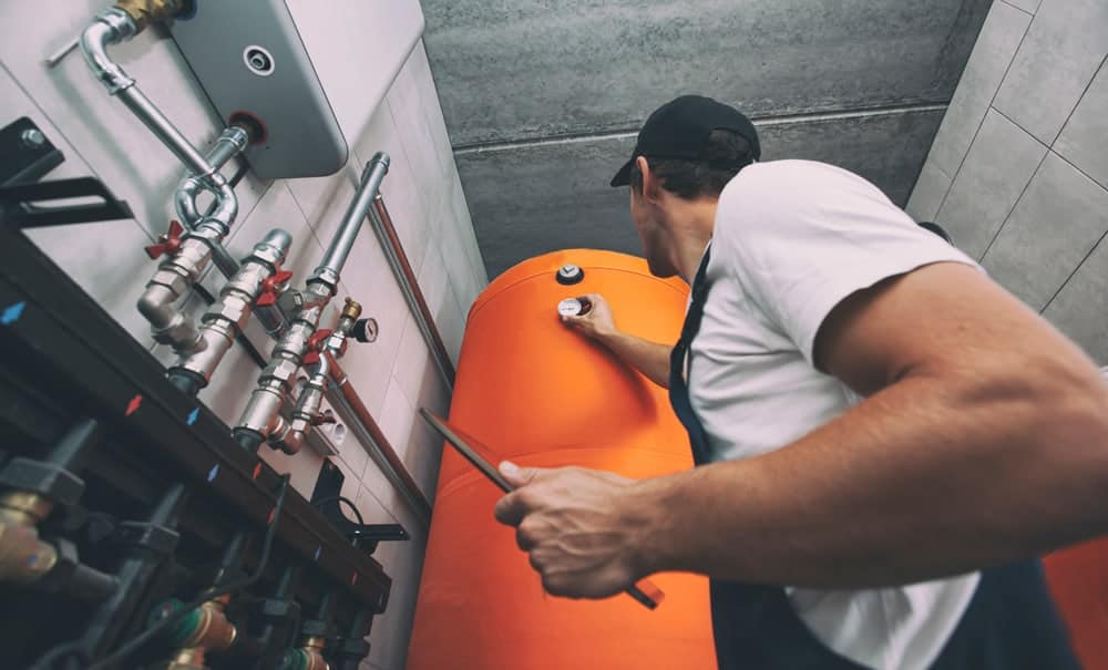 A worker in a white shirt and cap checks the pressure gauge on a large orange water heater tank in a NY utility room, showcasing expertise typical of a Plumbing Company New York County & Staten Island trusts for quality service.