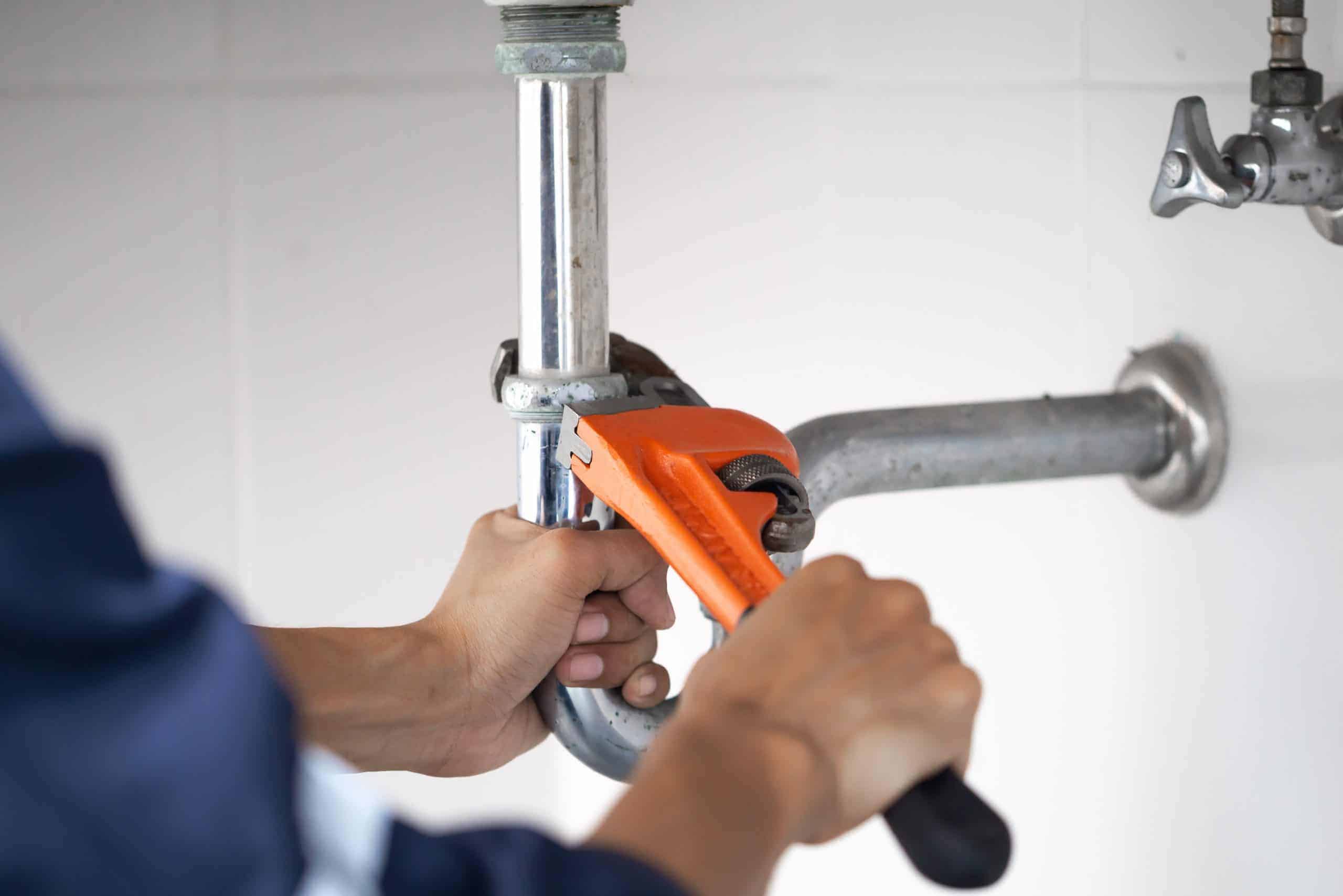 A person uses an orange pipe wrench to tighten or loosen a metal pipe under a sink, suggesting plumbing repair or maintenance work by a Plumbing Company in New York County & Staten Island, NY.