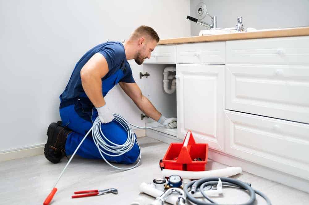 A plumber from a Plumbing Company New York County & Staten Island, NY, wearing blue overalls, kneels on the floor to repair pipes under a white bathroom sink, holding a coiled cable. Plumbing tools and equipment are scattered nearby.