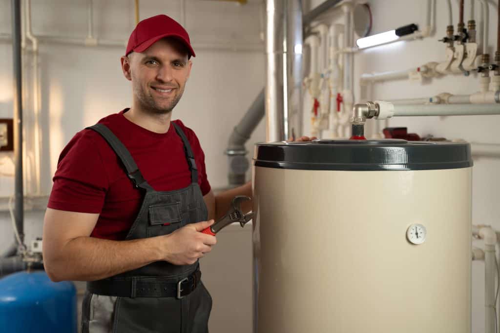 A plumber in a red cap and overalls smiles while holding a wrench next to a large water heater in a utility room with pipes and valves.