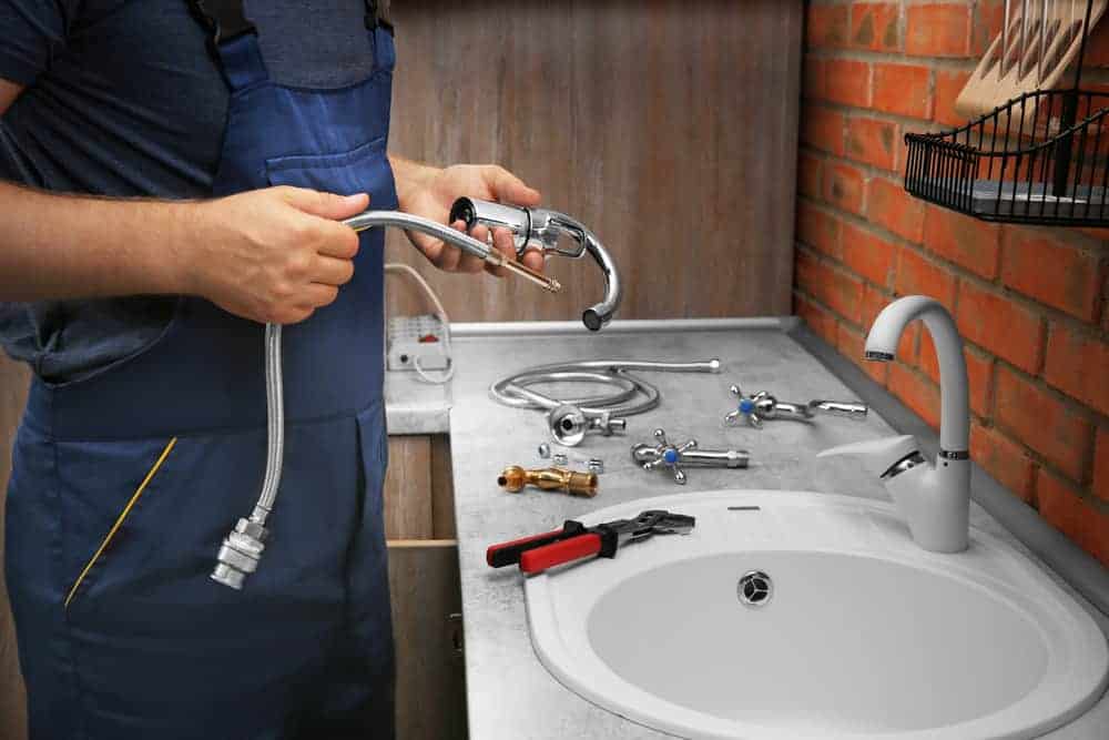 A plumber in blue overalls from a Plumbing Company New York County & Staten Island holds a faucet and hose above a bathroom sink, with various plumbing tools and parts laid out on the countertop. A modern faucet is already installed on the sink.