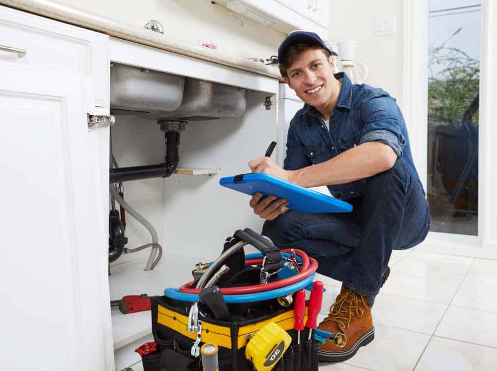 A smiling plumber from a top Plumbing Company New York County & Staten Island, NY kneels by an open sink cabinet, holding a clipboard. A tool bag with various tools sits on the floor beside him in a bright, modern kitchen.