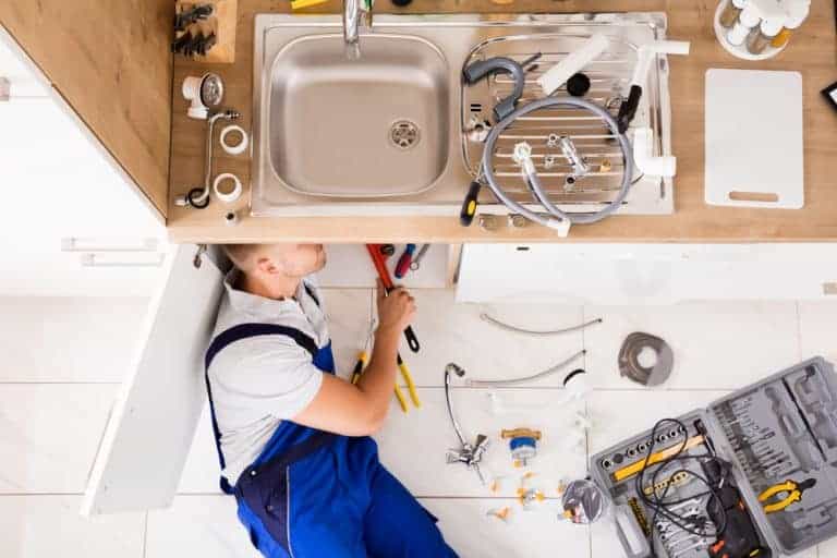 A plumber from a Plumbing Company New York County & Staten Island, NY lies under a kitchen sink, fixing pipes with tools scattered on the floor and countertop, including wrenches, hoses, and various plumbing parts.