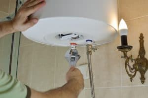 A person uses a wrench to adjust a valve underneath a white water heater mounted on a bathroom wall, with beige tiles and a decorative wall lamp nearby.