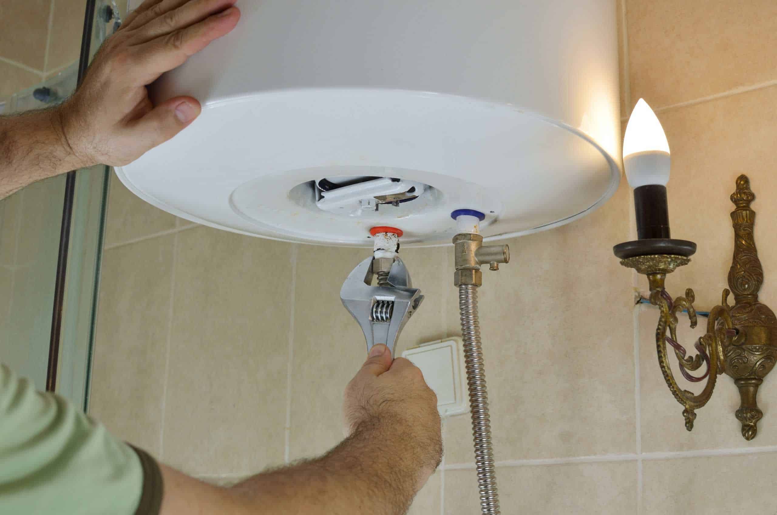 A person uses a wrench to adjust a valve on the underside of a wall-mounted water heater in a tiled NY bathroom, with a light fixture visible nearby&mdash;a common service by Plumbing Company New York County & Staten Island.