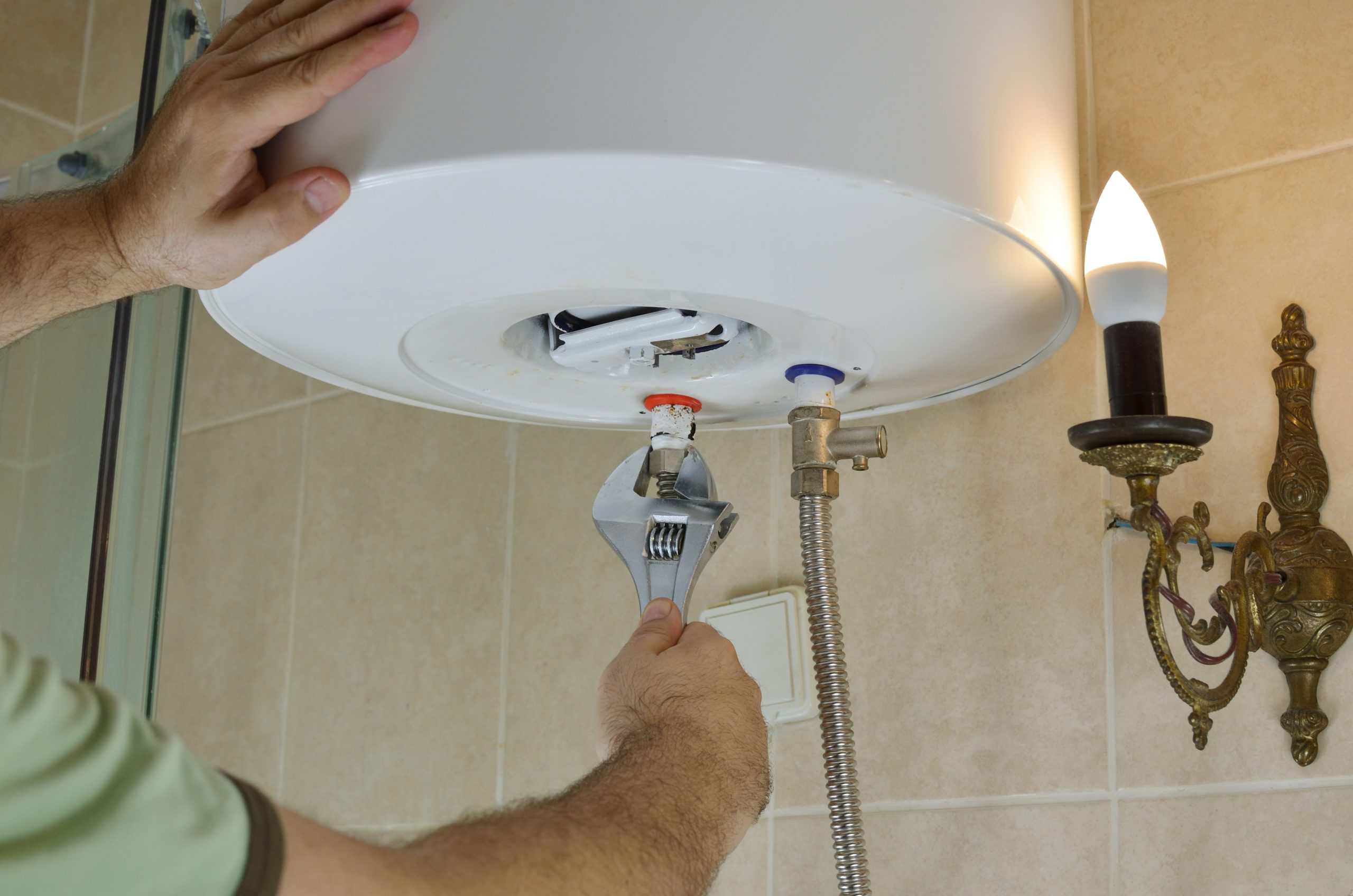 A person uses a wrench to adjust a valve underneath a white water heater mounted on a bathroom wall, with beige tiles and a decorative wall lamp nearby.