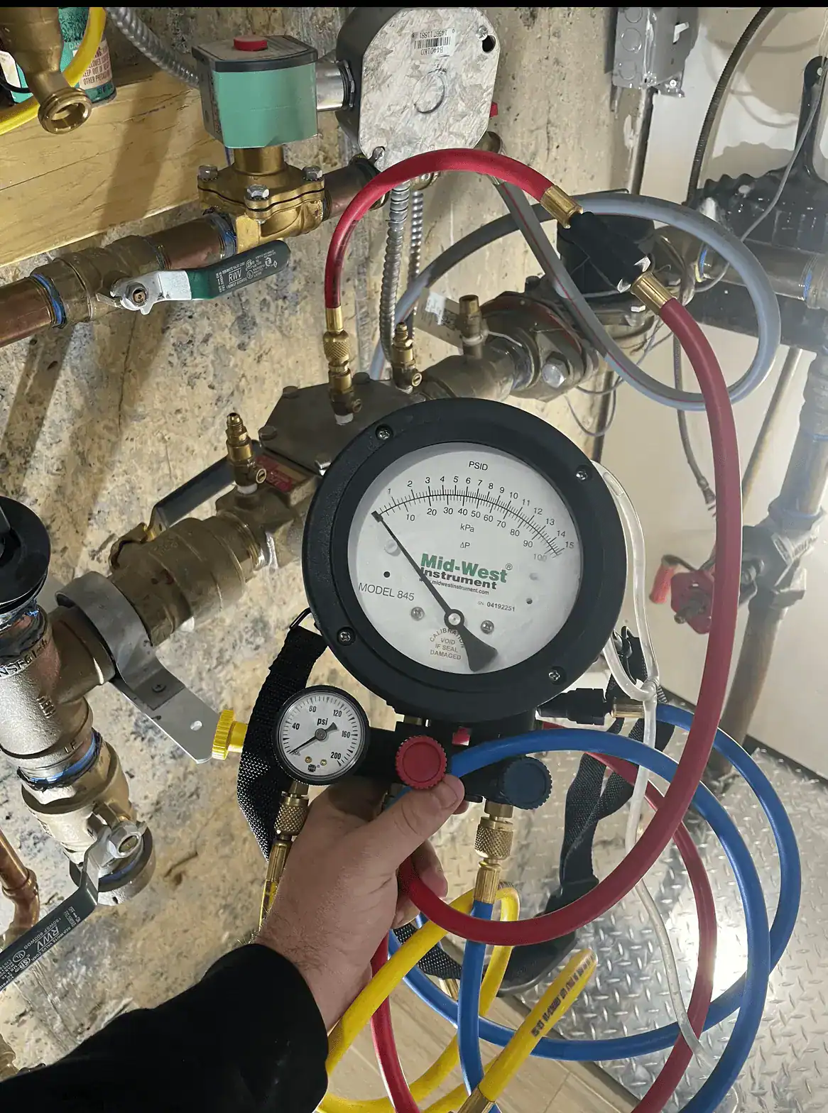A hand holds a Mid-West differential pressure gauge connected to colorful tubes and pipes on a wall, likely measuring system pressure in a mechanical or industrial setting.